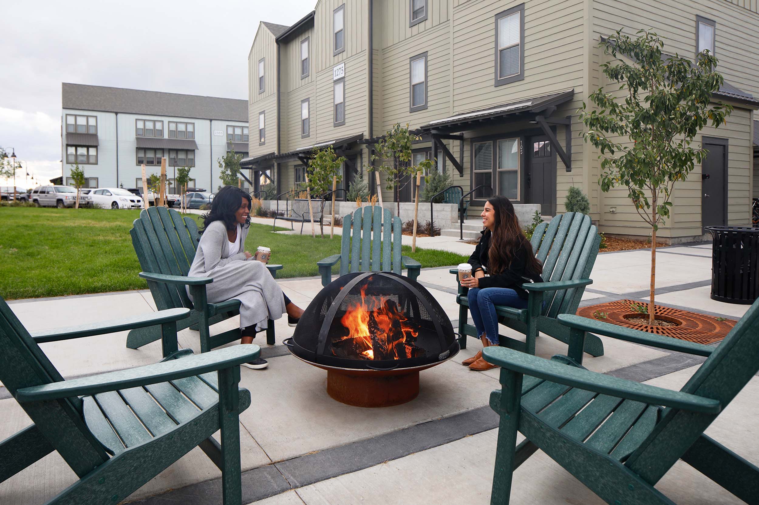 Friends sitting in green deck chairs next tof firepit outside apartment buildings